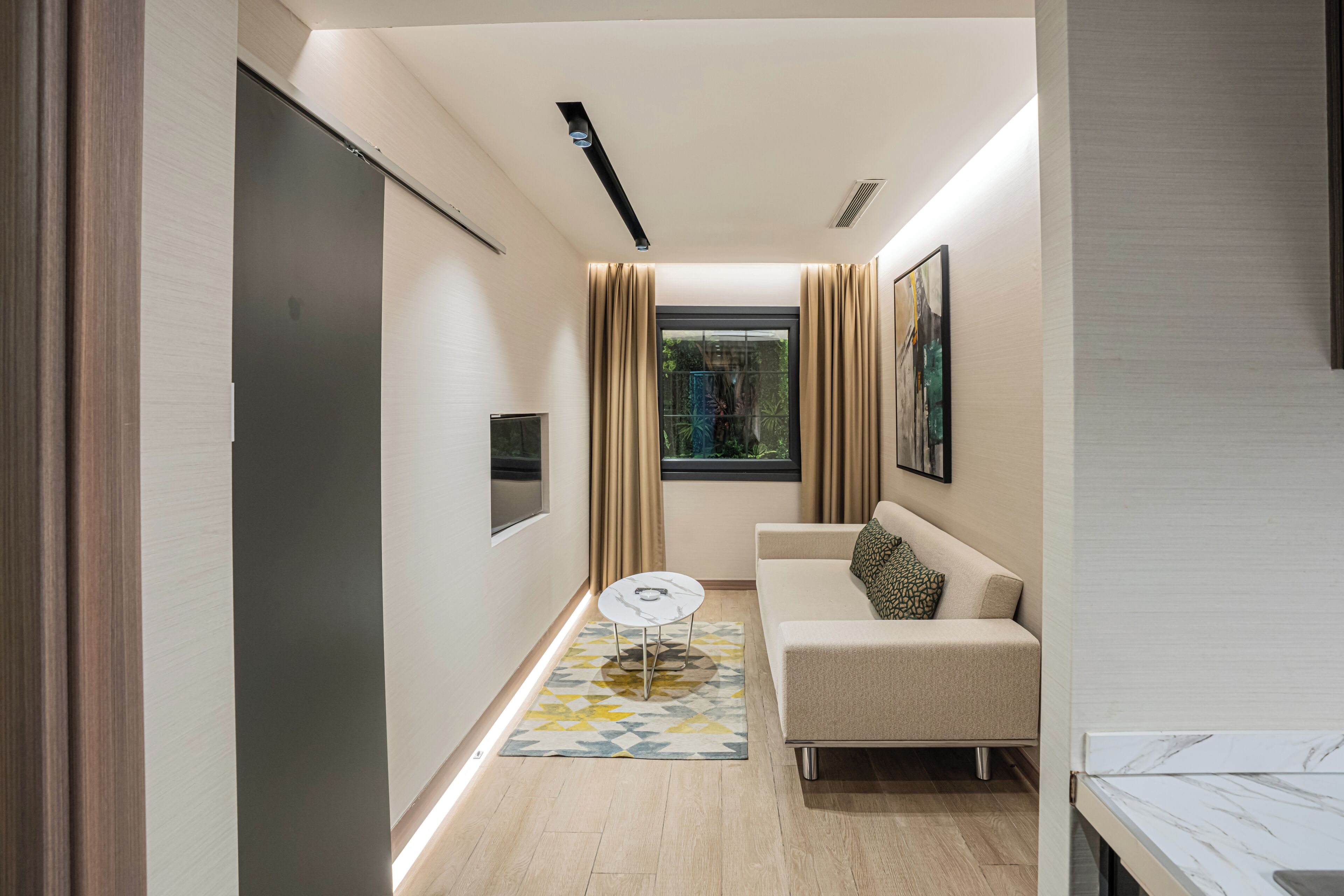 A modern hotel room with a sofa, a small round table, a TV, and a window with curtains.
