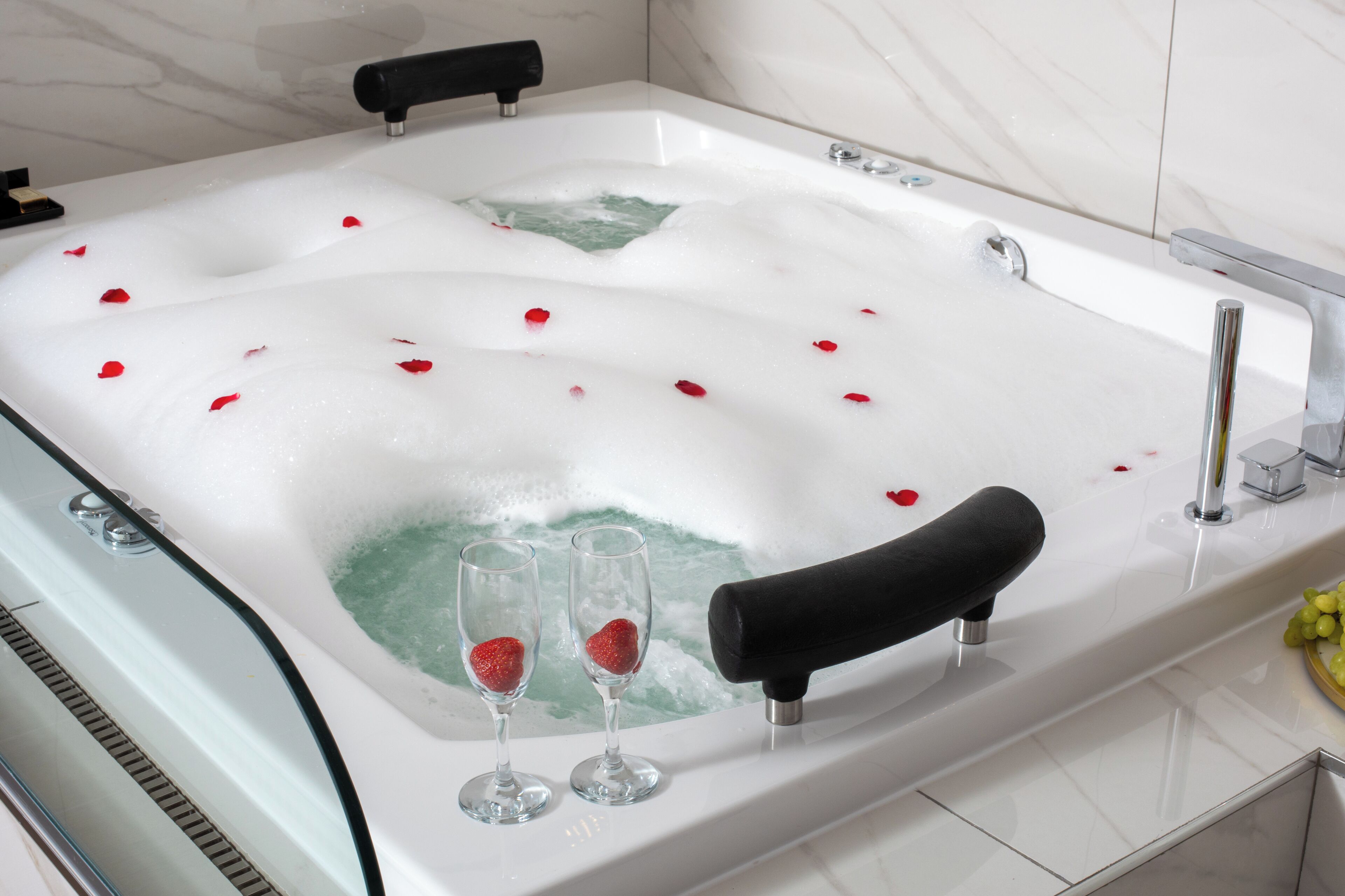 A bathtub filled with bubbles and rose petals, with two champagne flutes and a black bath mat.