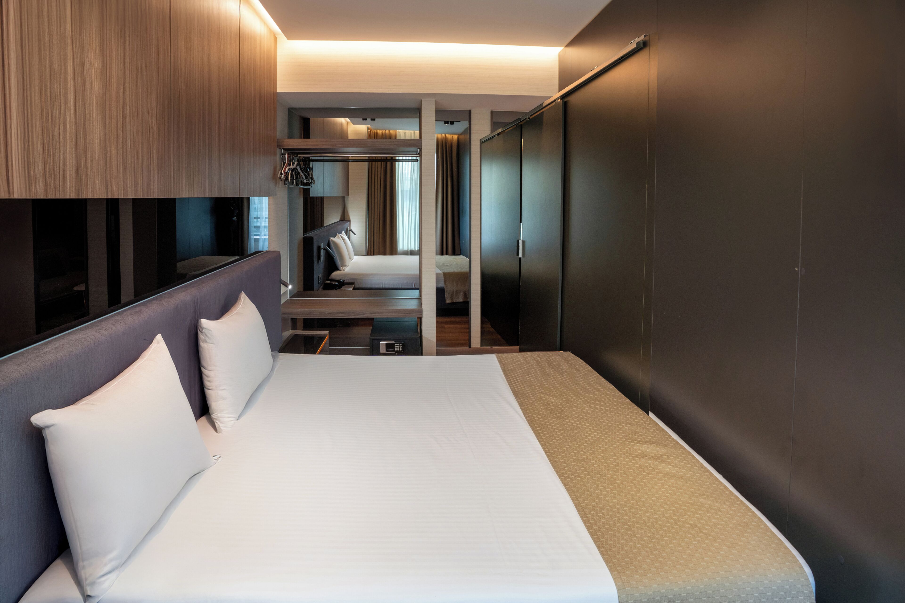 A modern hotel room with a large bed, a seating area with a sofa and chairs, a wardrobe, and a small desk with a lamp.