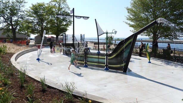 Zona de juegos acuáticos al aire libre con tobogán y chorros de agua, rodeada por un camino pavimentado y árboles.