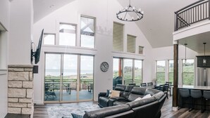 A spacious living area with a large black leather sofa, a stone fireplace, and a view of a patio with outdoor furniture. - Unobstructed Lake Views! Bunk Room w/ Video Games! Ping Pong & Foosball! Extra Fridge & Freezer Space! (Fish Haven)