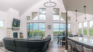 A spacious living area with a stone fireplace, a large black leather sofa, a dining table with chairs, and a kitchen island. - Unobstructed Lake Views! Bunk Room w/ Video Games! Ping Pong & Foosball! Extra Fridge & Freezer Space! (Fish Haven)