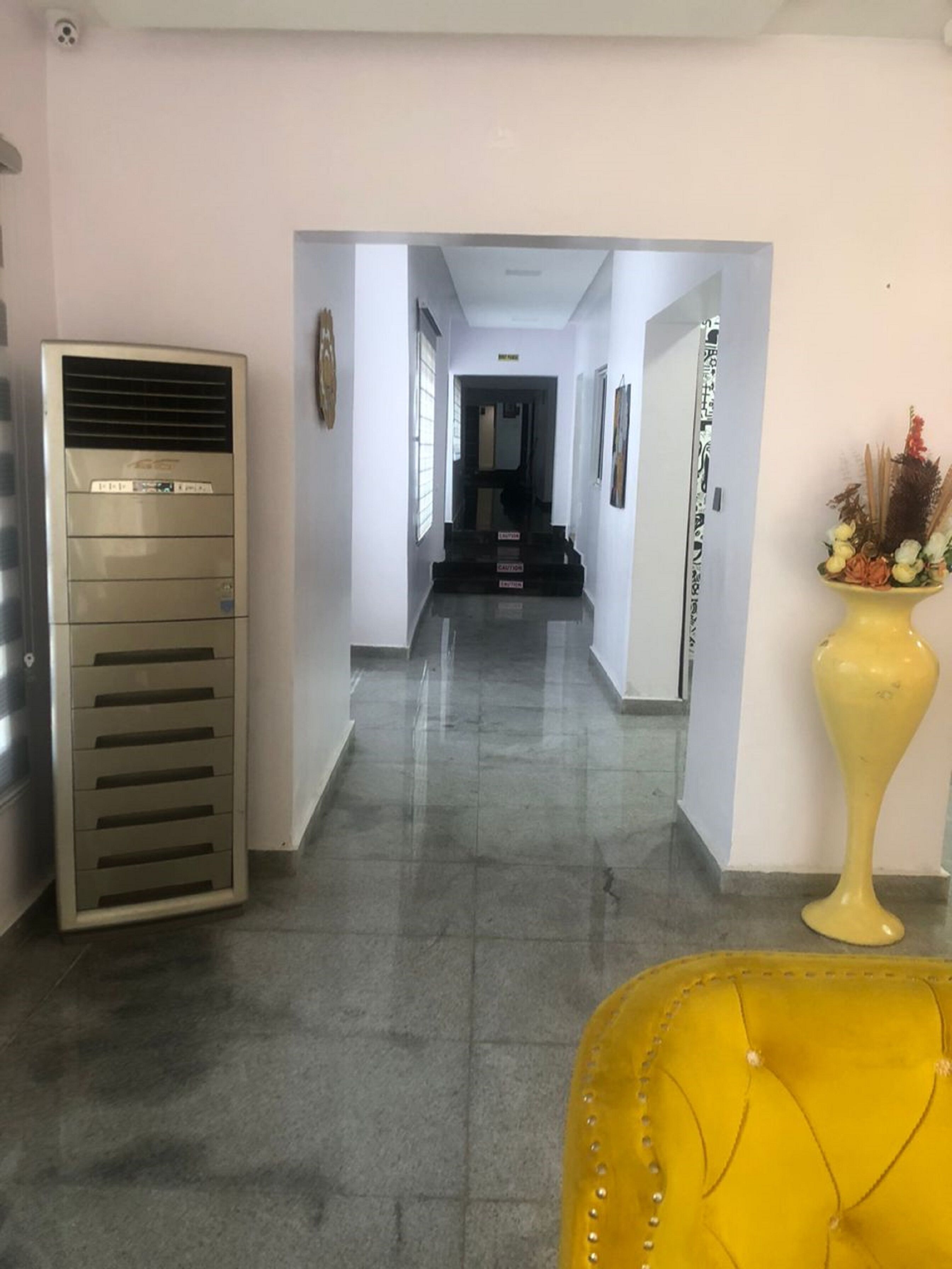 A hallway with a yellow chair, a large air conditioning unit, and a yellow vase with flowers.