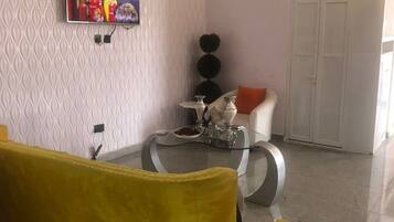 Ein Wohnzimmer mit gelber Couch, einem an der Wand befestigten Fernseher, einem dekorativen Tisch mit Vase und Krug sowie einem Kleiderschrank.