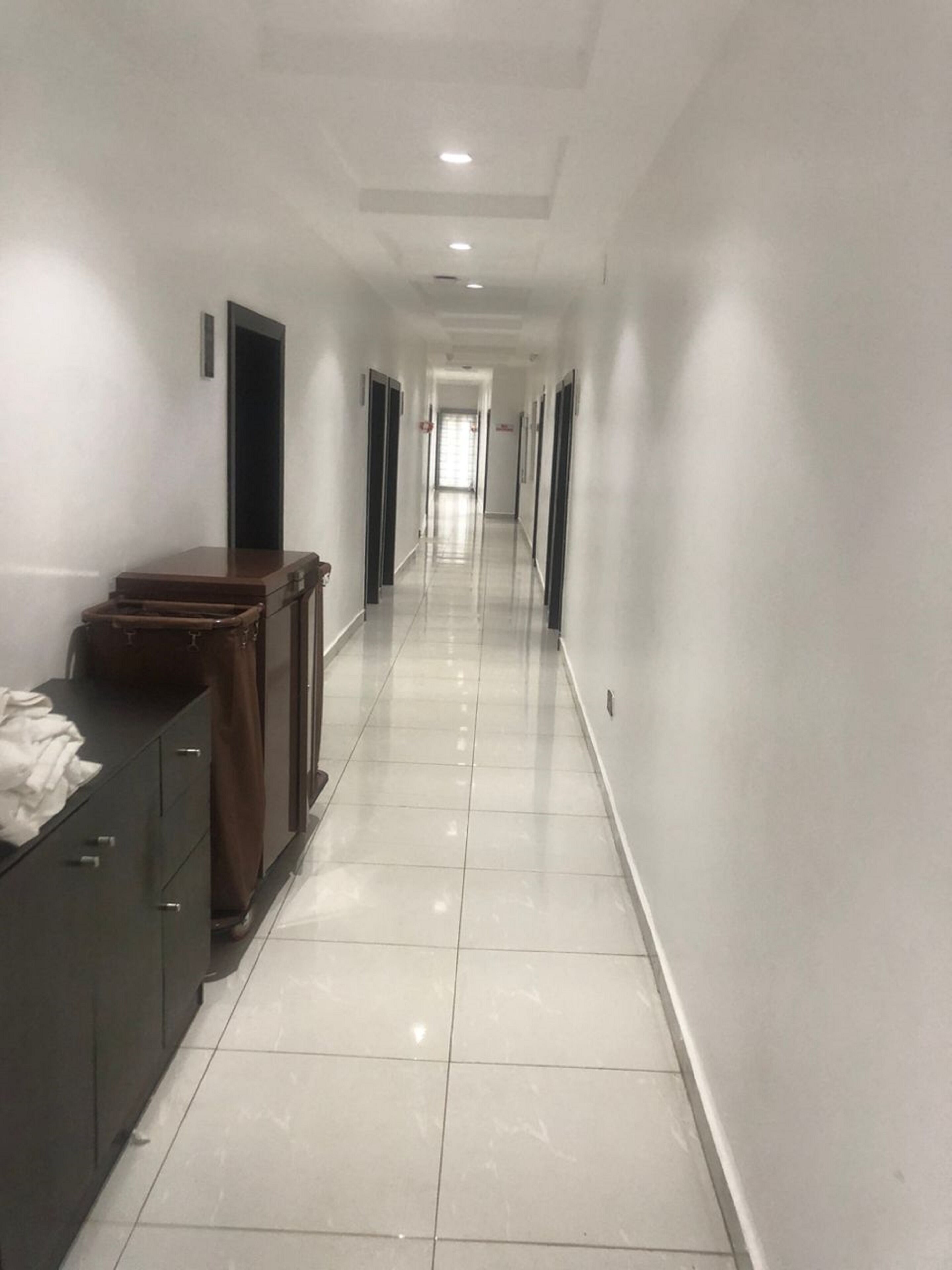 A long, narrow hallway with white walls and a tiled floor, leading to a door at the end.