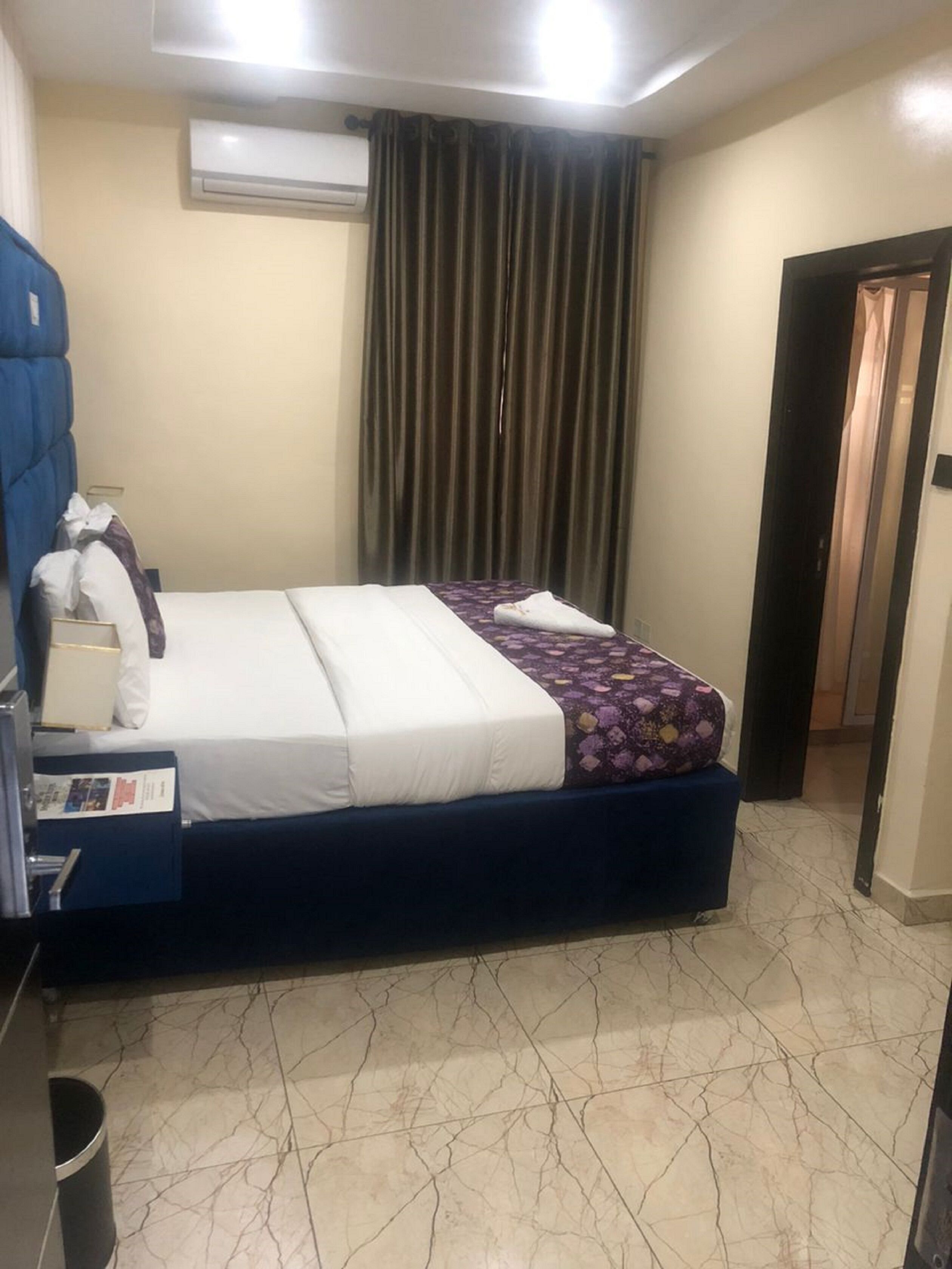 A hotel room with a bed, a desk with a chair, an air conditioning unit, and a door.