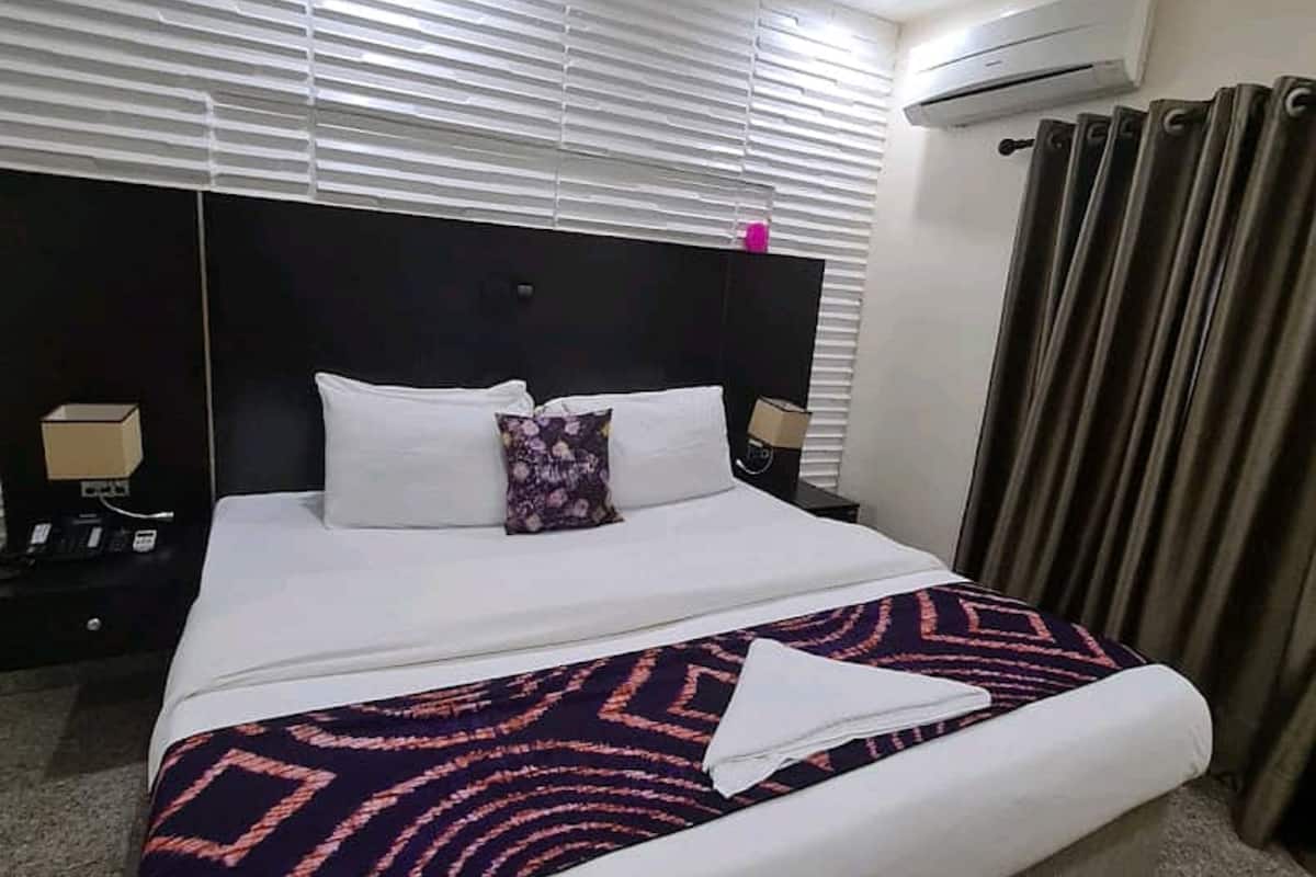 Une chambre d’hôtel avec un grand lit, une tête de lit sombre, du linge de lit blanc, un plaid à motifs et une table de chevet avec une lampe.