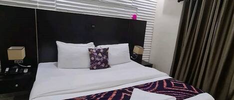 A hotel room with a large bed, a dark headboard, white bedding, a patterned throw blanket, and a nightstand with a lamp.