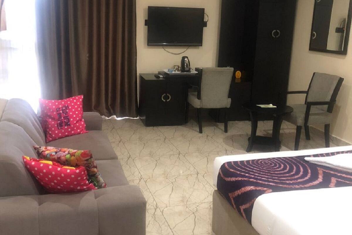 Une chambre d’hôtel avec un canapé gris, un lit recouvert d’une couvre-lit à motifs, un bureau avec une chaise, un téléviseur à écran plat, une armoire et un grand miroir.