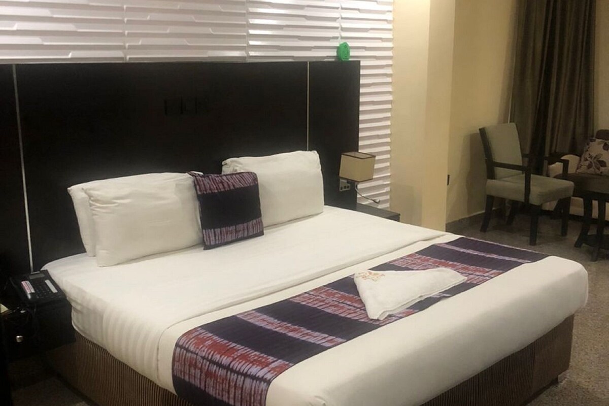 Habitación de hotel con una cama grande, cabecera, mesita de noche con lámpara, una silla y una pequeña mesa con otra silla.