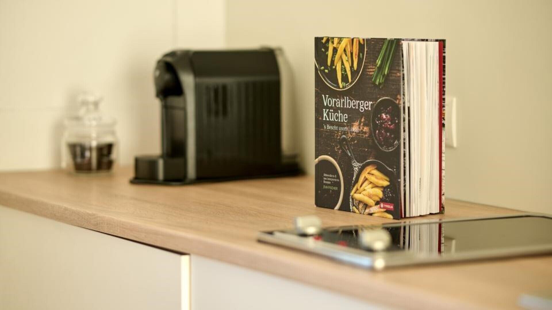 A kitchen counter with a coffee maker, a cookbook titled 'Vorarlberger Küche', and a smartphone.