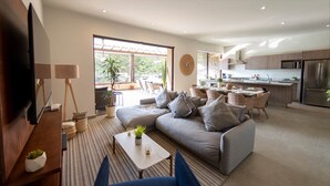 A modern living room with a sofa, coffee table, and TV. There is a kitchen area with a refrigerator and cabinets.