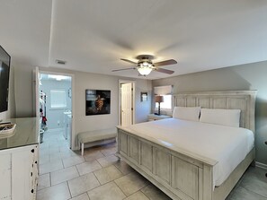 A bedroom with a large bed, a ceiling fan, a bench, a TV, and a painting on the wall.