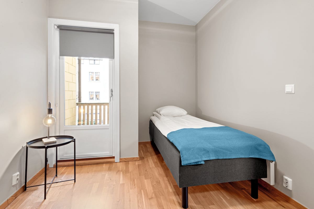 A small, neatly arranged bedroom with a single bed, a small table, and a door leading to an outdoor balcony.