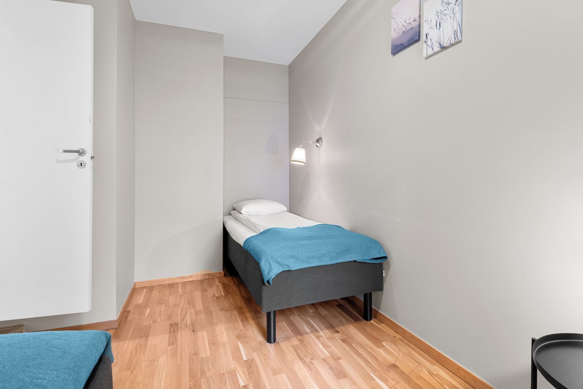 A small, modern bedroom with a single bed, a bedside table, a wall-mounted light, and a door.