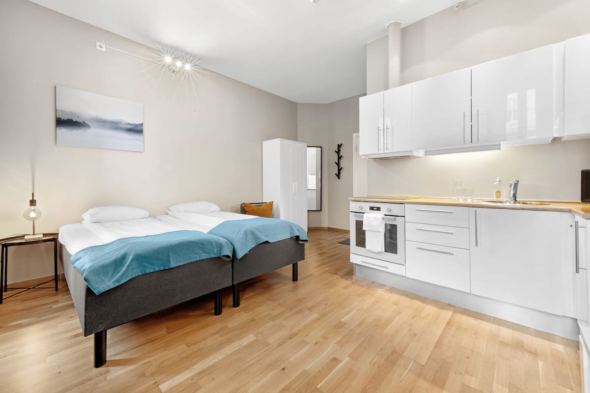 A modern bedroom with a bed, bedside table, and a nightstand. Adjacent to the bedroom is a kitchen with white cabinets and a wooden countertop.