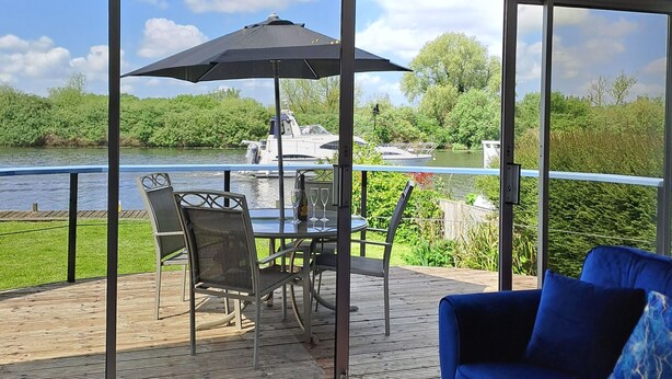 Ein Zimmer mit großem Fenster und Blick auf einen Fluss, eine Terrasse mit Gartenmöbeln und ein blaues Sofa.