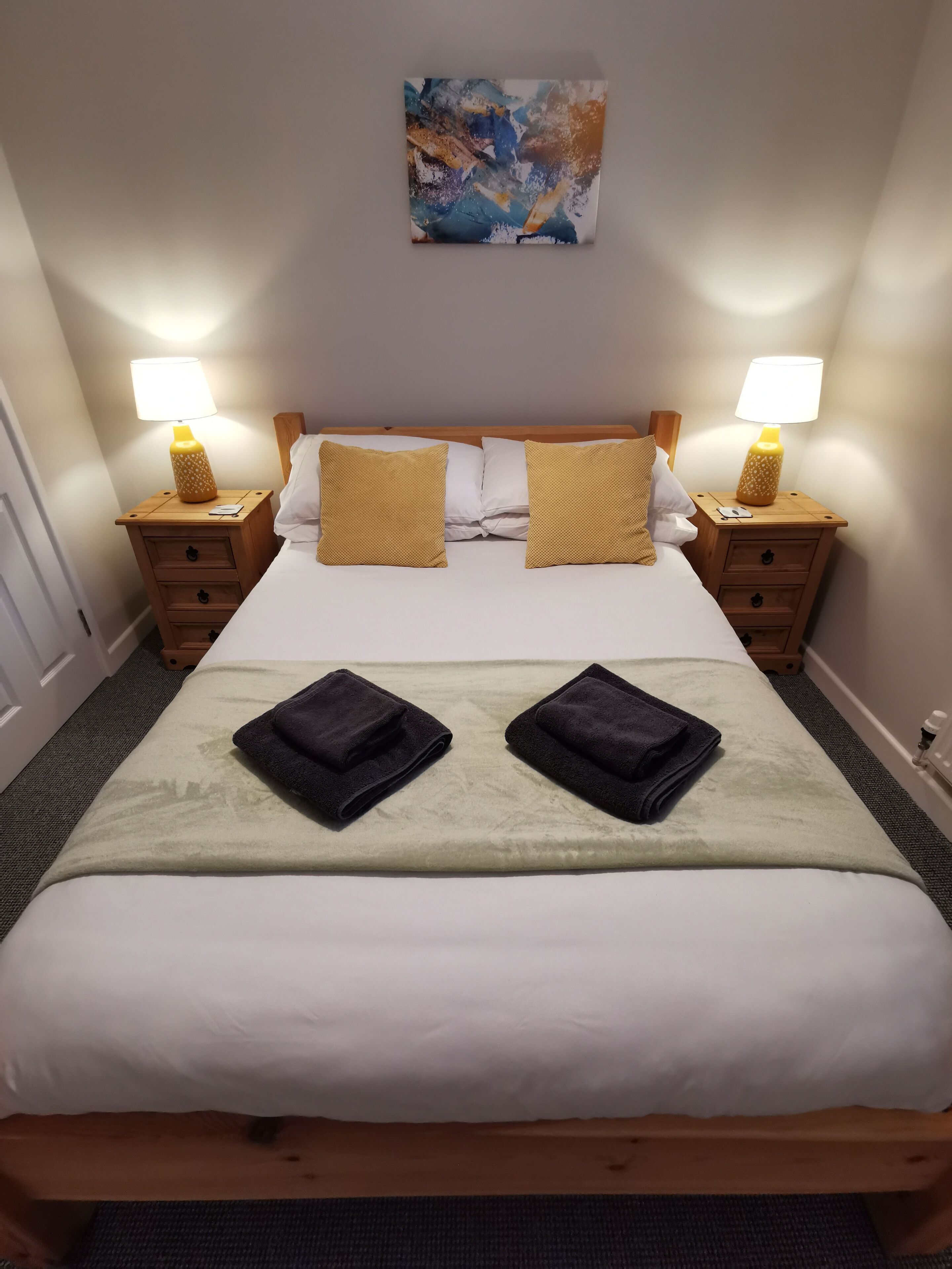 A neatly made bed with two pillows, a blanket, and two folded towels. There are two bedside tables with lamps, a wooden headboard, and a painting above the bed.