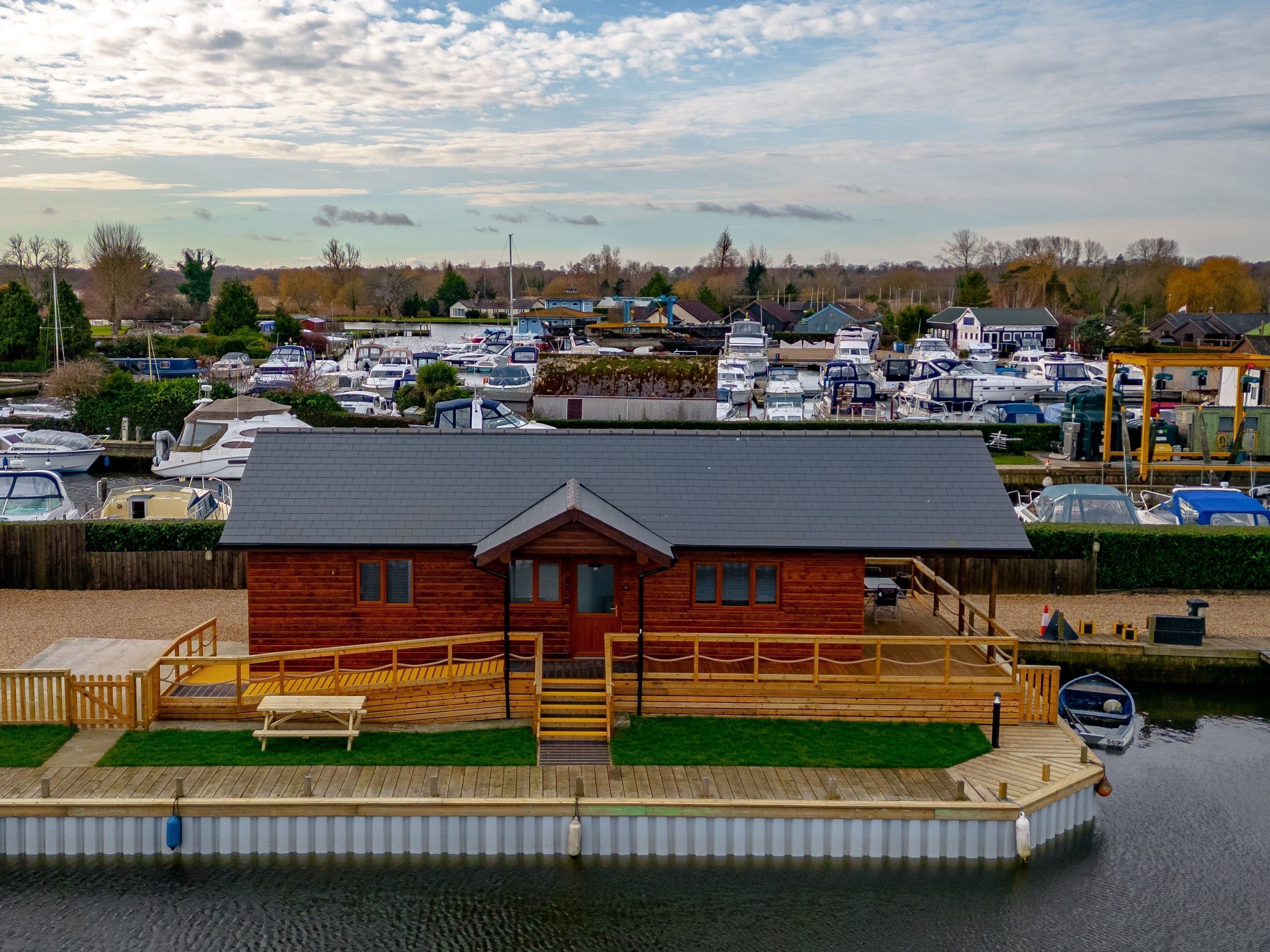 A wooden dock with a small house, a grassy area, and a marina with boats in the background.