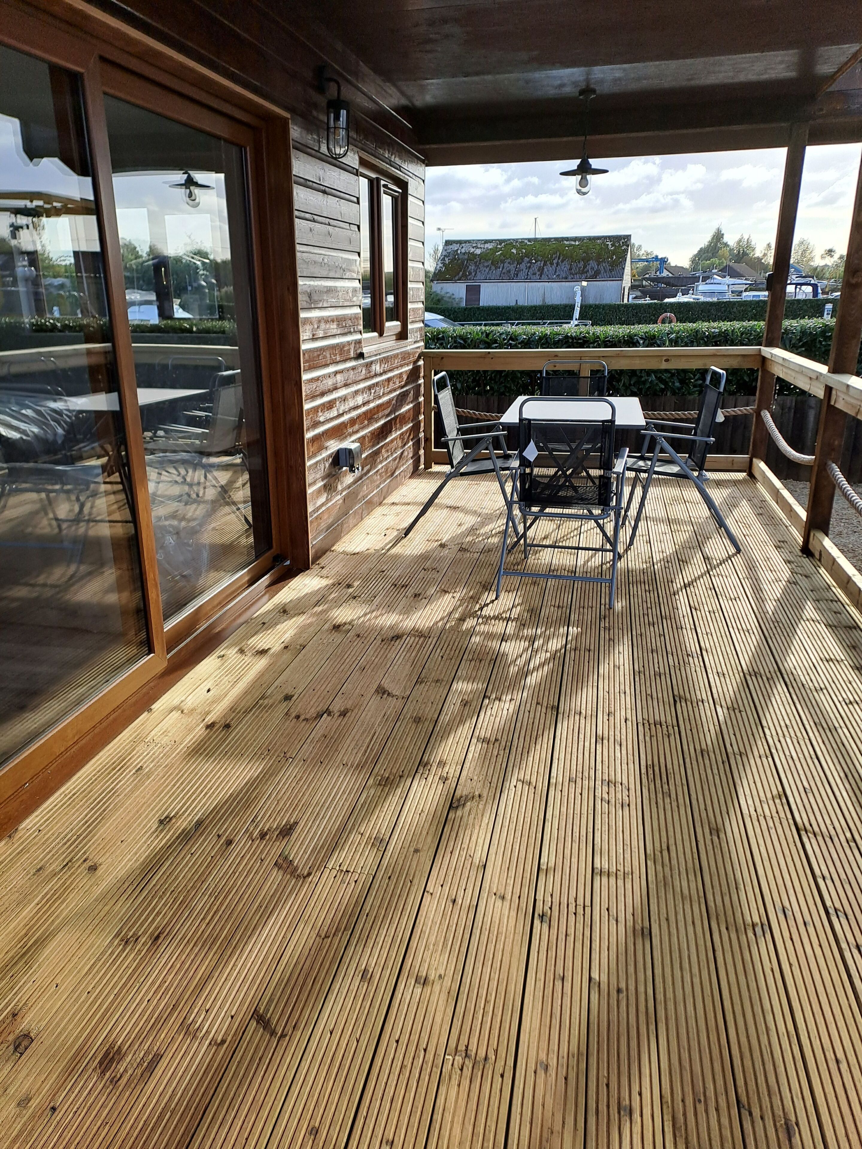 A wooden deck with a table and chairs, large sliding glass doors, and a view of a garden and buildings in the background.