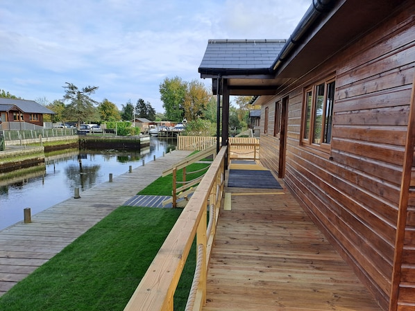 A wooden cabin with a dock extending into a calm waterway, surrounded by greenery and other cabins.