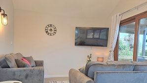Un salon avec deux canapés gris, une petite table ronde, une horloge murale, un téléviseur à écran plat et une fenêtre avec des rideaux.