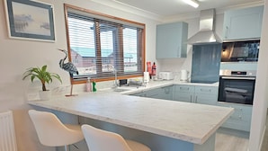 Une cuisine moderne dotée d’un plan de travail en marbre, de deux tabourets de bar blancs, d’une fenêtre avec des stores et d’un four Bosch.