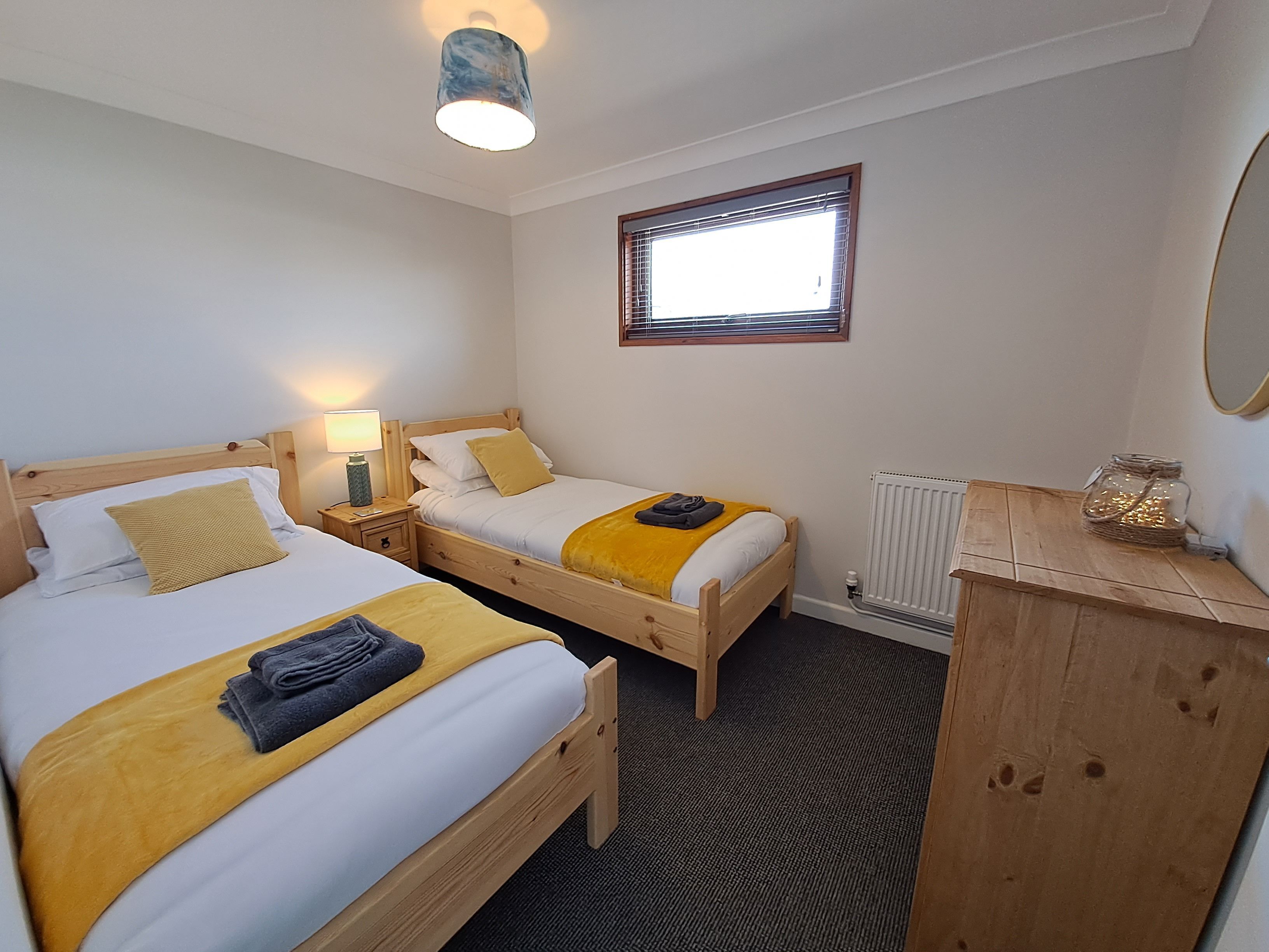 A room with two wooden beds, a small table, a lamp, a radiator, and a window with blinds.