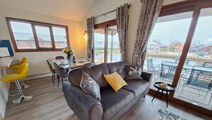 A living room with a grey sofa, yellow chair, dining table, and large windows with curtains. - River Breeze - Waterside, Norfolk Broads, sleeps 6 in 3 bedrooms (Brundall)