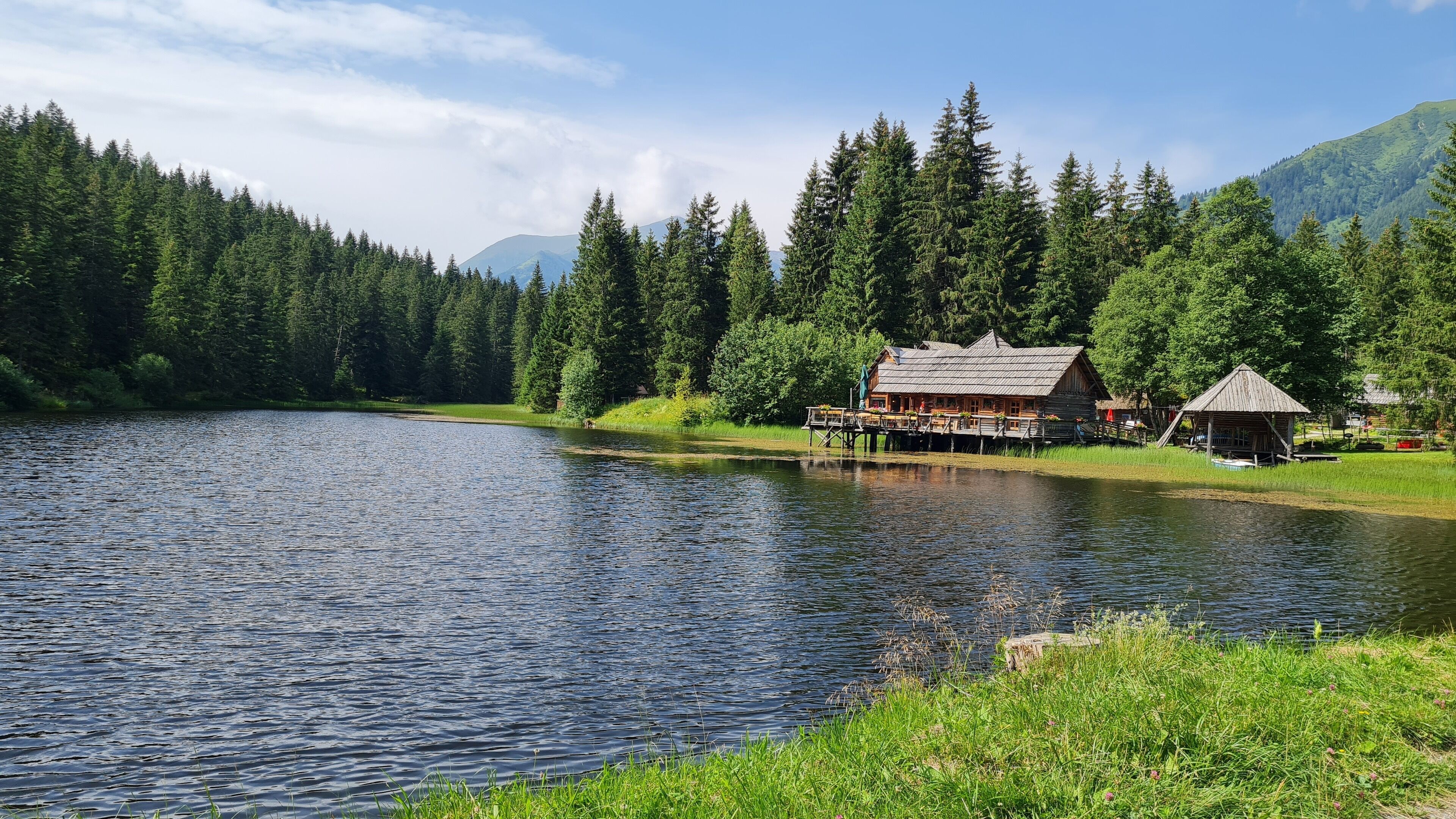 A lakeside cabin with a wooden deck and a small shed, surrounded by dense forest and mountains.