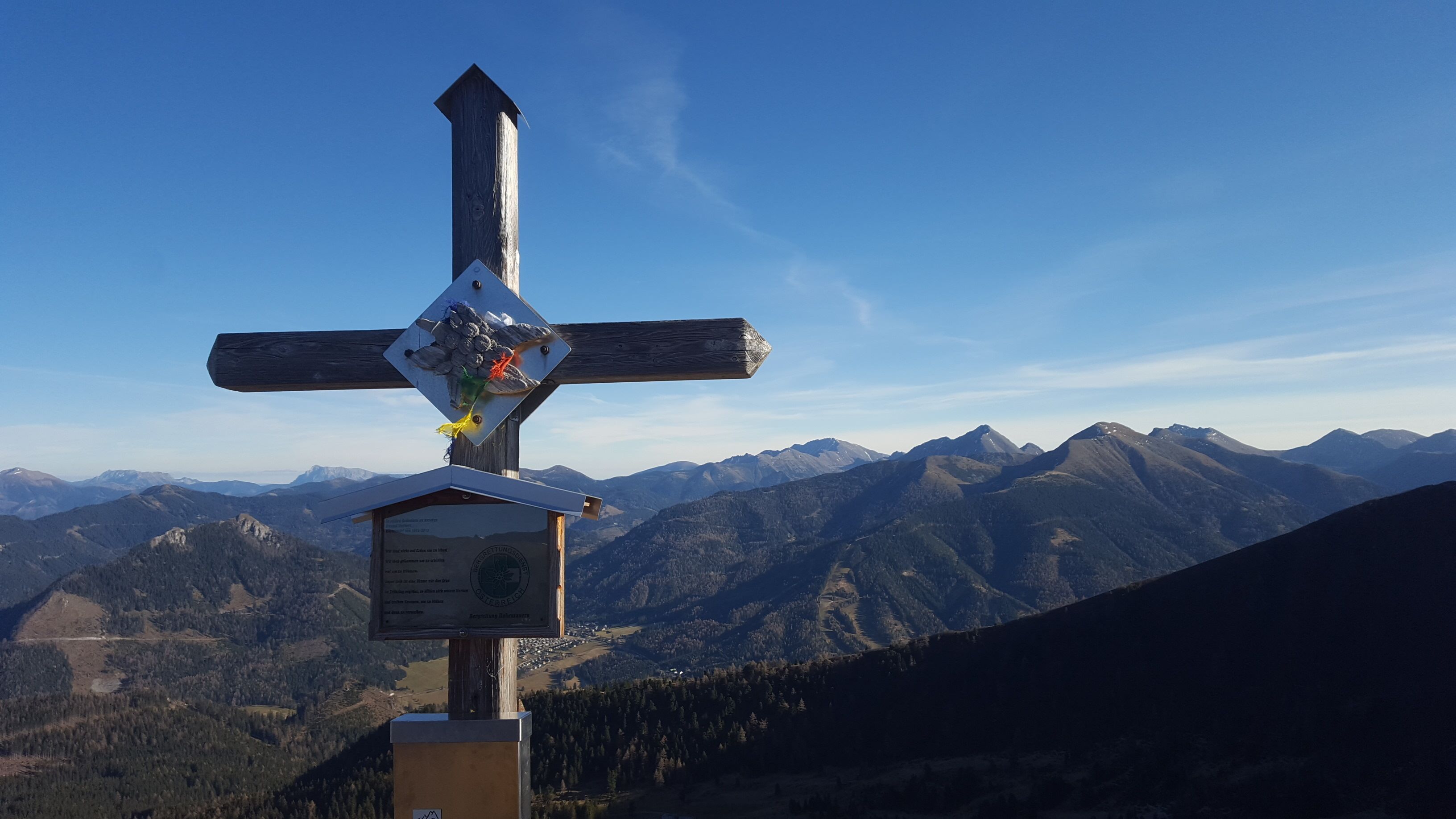 Ein hölzernes Kreuz mit einem Schild und einer kleinen Struktur an der Basis, vor einer Berglandschaft.