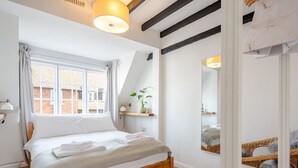 A bedroom with a wooden bed, white bedding, a window with curtains, a bedside lamp, a mirror, and a plant on a shelf. - The Hay Loft - Pass the Keys (Canterbury)