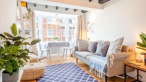 A cozy living room with a sofa, a coffee table, and a dining area with chairs. There is a large window providing natural light and a view of the outside. - The Hay Loft - Pass the Keys (Canterbury)