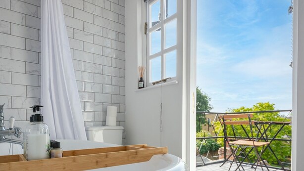 A bathroom with a bathtub, a toilet, and a window leading to a balcony with outdoor furniture. - The Hay Loft - Pass the Keys (Canterbury)