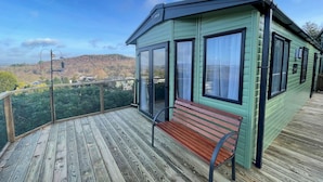 A wooden deck with a bench, a green building with large windows, and a view of a forested area. - Beautiful 2BR In Kippford With Incredible Views - Pass the Keys (Dalbeattie)