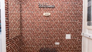 A room with a brick wall, a 'Welcome' sign, a 'Sweet Home' sign, a black mat, and a white door.