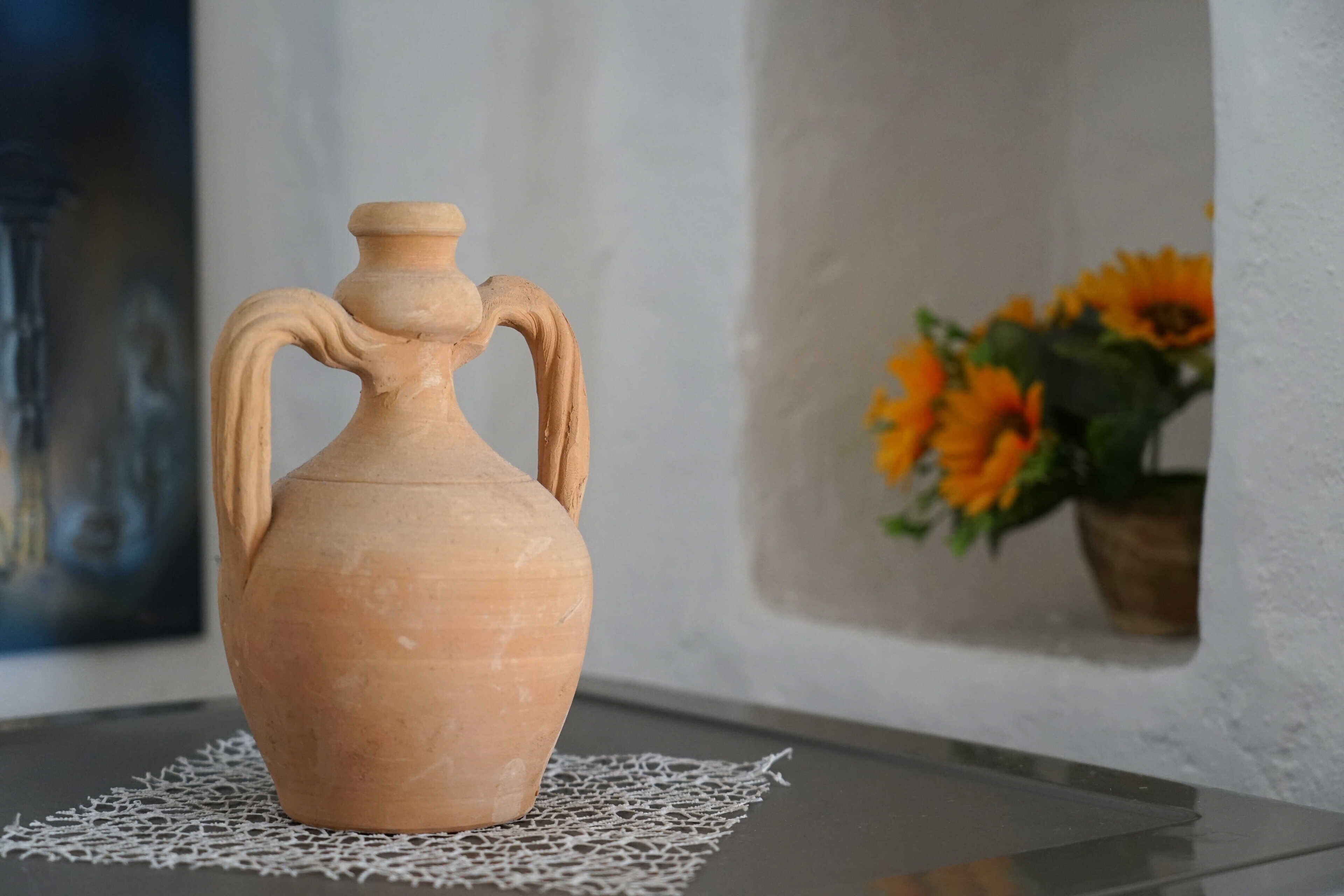 Una jarra de cerámica con asas sobre una mesa con un mantel de encaje, junto a una pared con un arreglo floral.