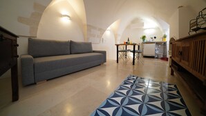 Una sala de estar acogedora con un sofá gris, un tapete estampado y una zona de cocina con una mesa y sillas.