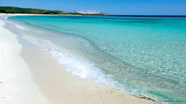 Una playa con agua turquesa cristalina, arena blanca y una colina distante.