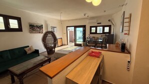 Una sala de estar con un sofá, un banco de madera, una mesa de comedor con sillas y una zona de cocina con mostrador y electrodomésticos.