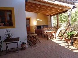 A covered patio with wooden furniture, a stone barbecue area, and potted plants.