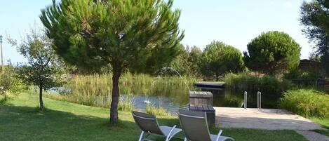 A serene outdoor area with a pond, two metal chairs, a wooden deck, and a tree.