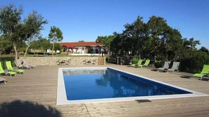 A rectangular outdoor swimming pool with lounge chairs and tables around it, a covered seating area, and trees in the background.