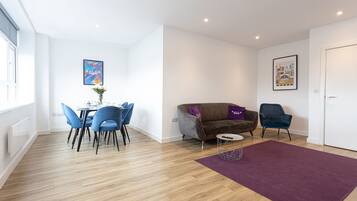 Una sala moderna con piso de madera, un tapete morado, un sofá marrón con un cojín morado, un sillón azul y un comedor con sillas y una mesa azules.