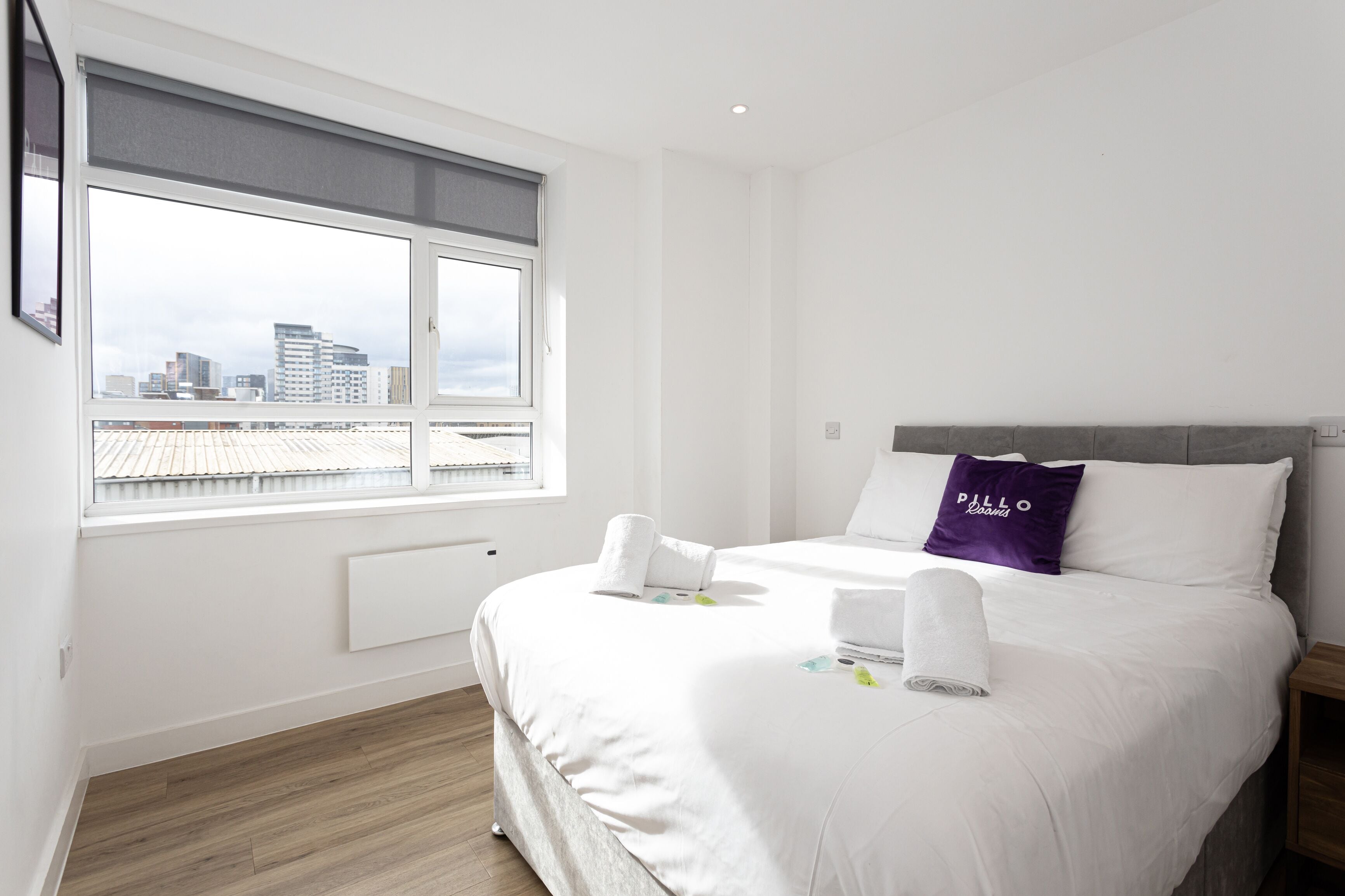 Une chambre d’hôtel avec une grande fenêtre, un lit avec une literie blanche, un oreiller violet et une vue sur le paysage urbain.