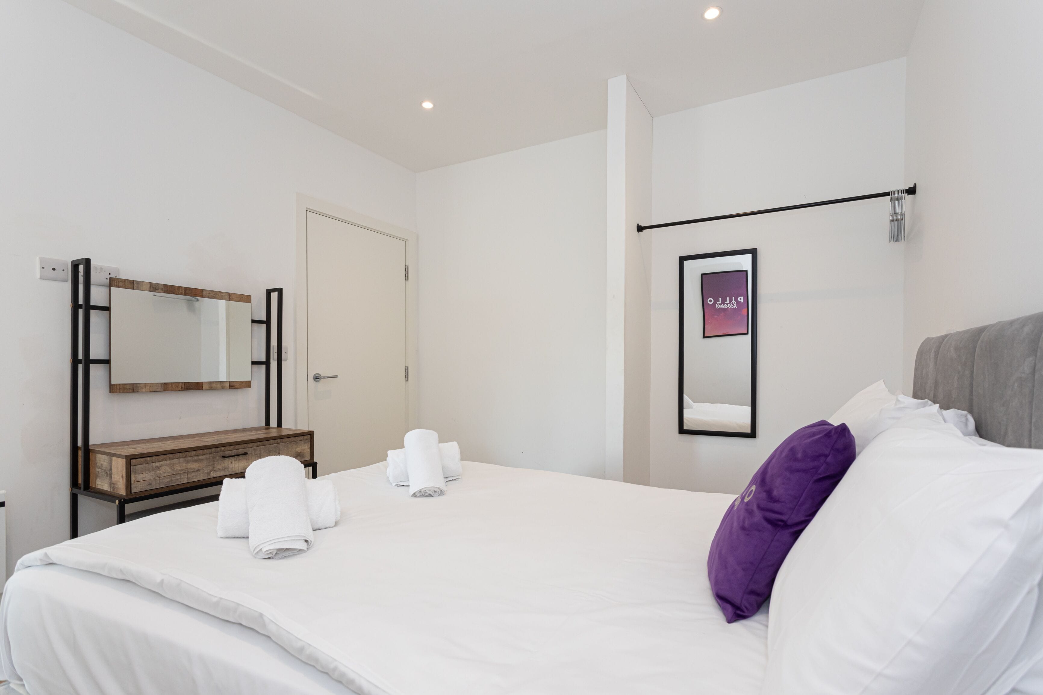 A hotel room with a bed, a wooden bedside table, a mirror, a towel rack, and a wall-mounted sign.