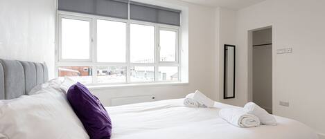 A hotel room with a large window, a bed with white linens, a purple pillow, and a mirror on the wall.