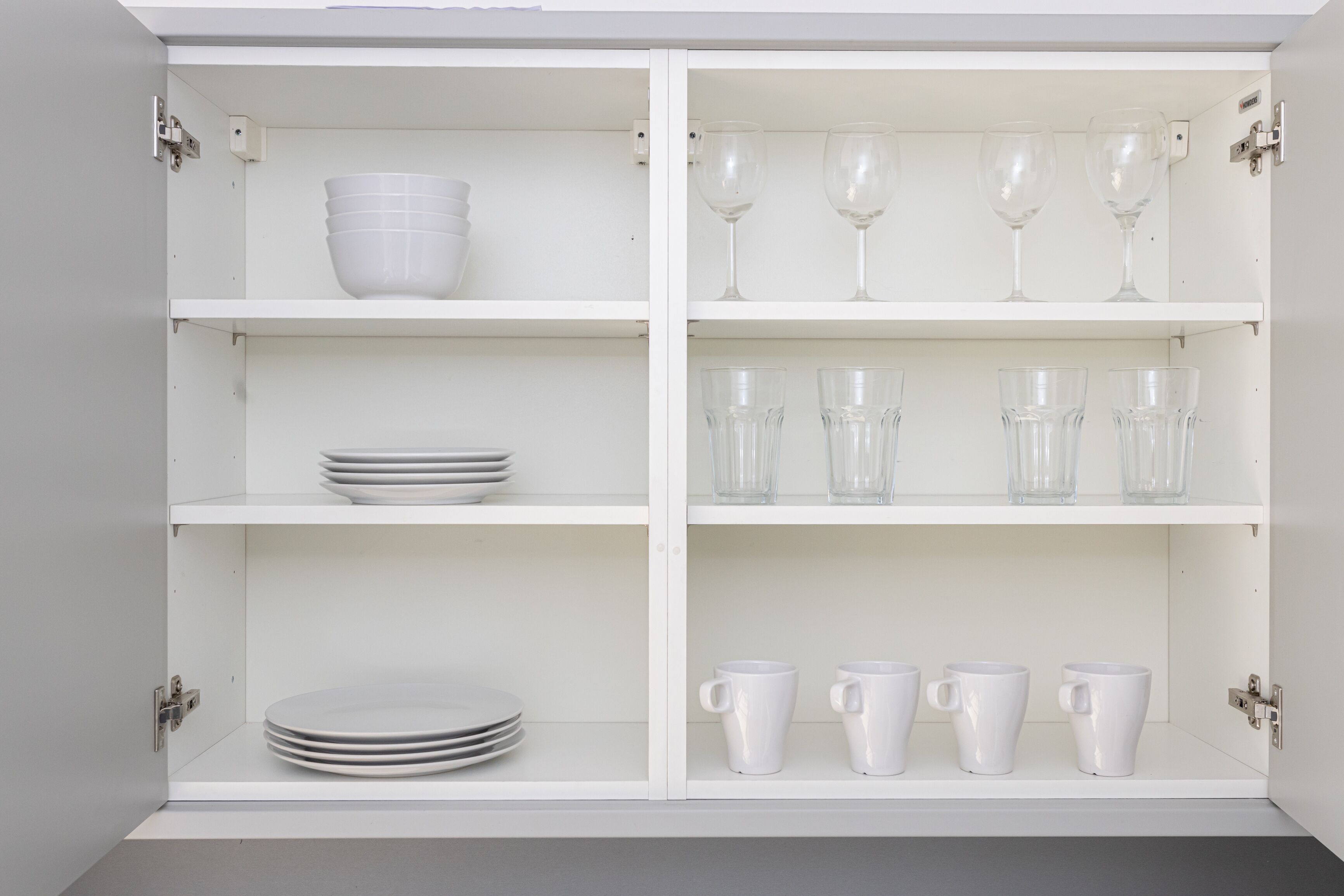 A white cabinet with four shelves, each holding different items: plates, wine glasses, and cups.
