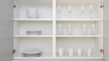 A white cabinet with four shelves, each holding different items: plates, wine glasses, and cups.