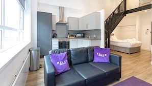 A modern apartment with a kitchen, a black leather sofa with purple pillows, and a staircase leading to a loft bedroom. - Pillo Rooms Apartments- Manchester Arena (Manchester)